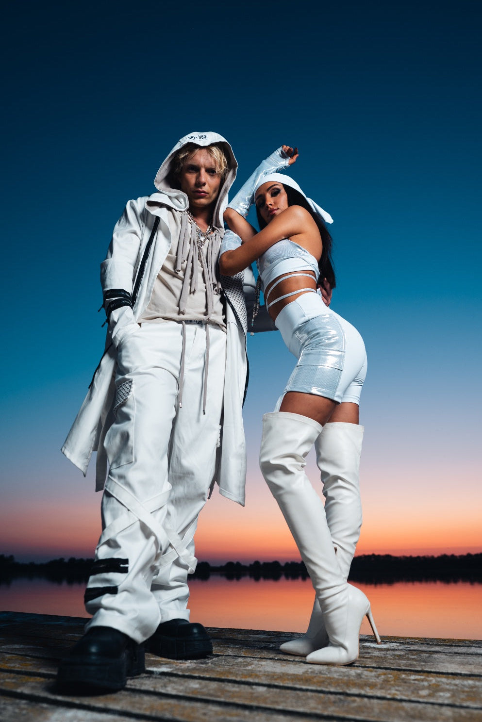 Two people in futuristic outfits standing on a dock with a sunset in the background