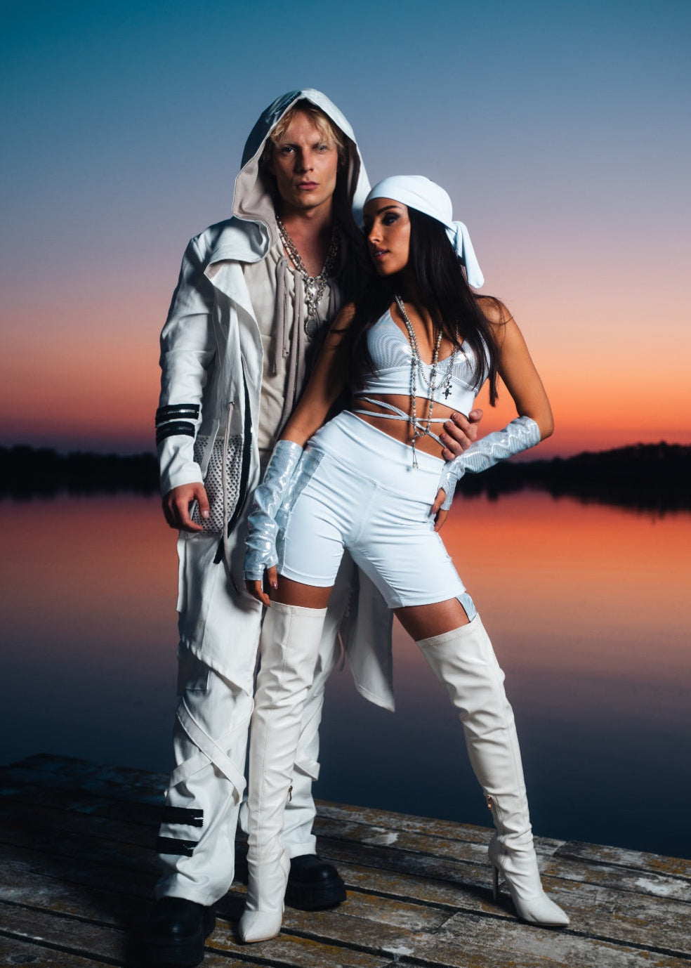 Two people in futuristic outfits standing on a dock at sunset.