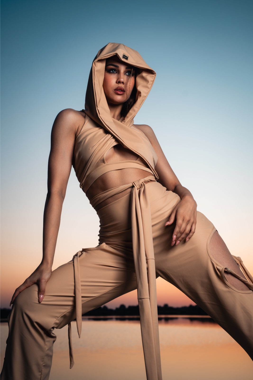 Person wearing a beige hooded outfit sitting by a body of water with a sunset sky.
