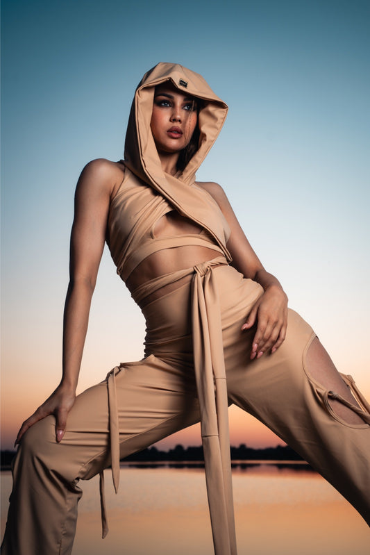 Person wearing a beige hooded outfit sitting by a body of water with a sunset sky.