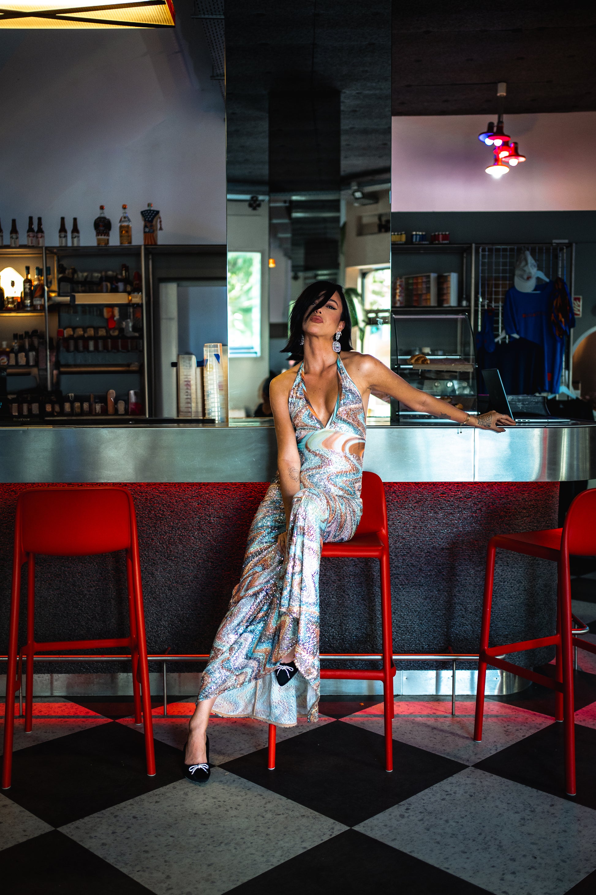 Woman sitting on a red bar stool in a bar setting with a checkered floor.