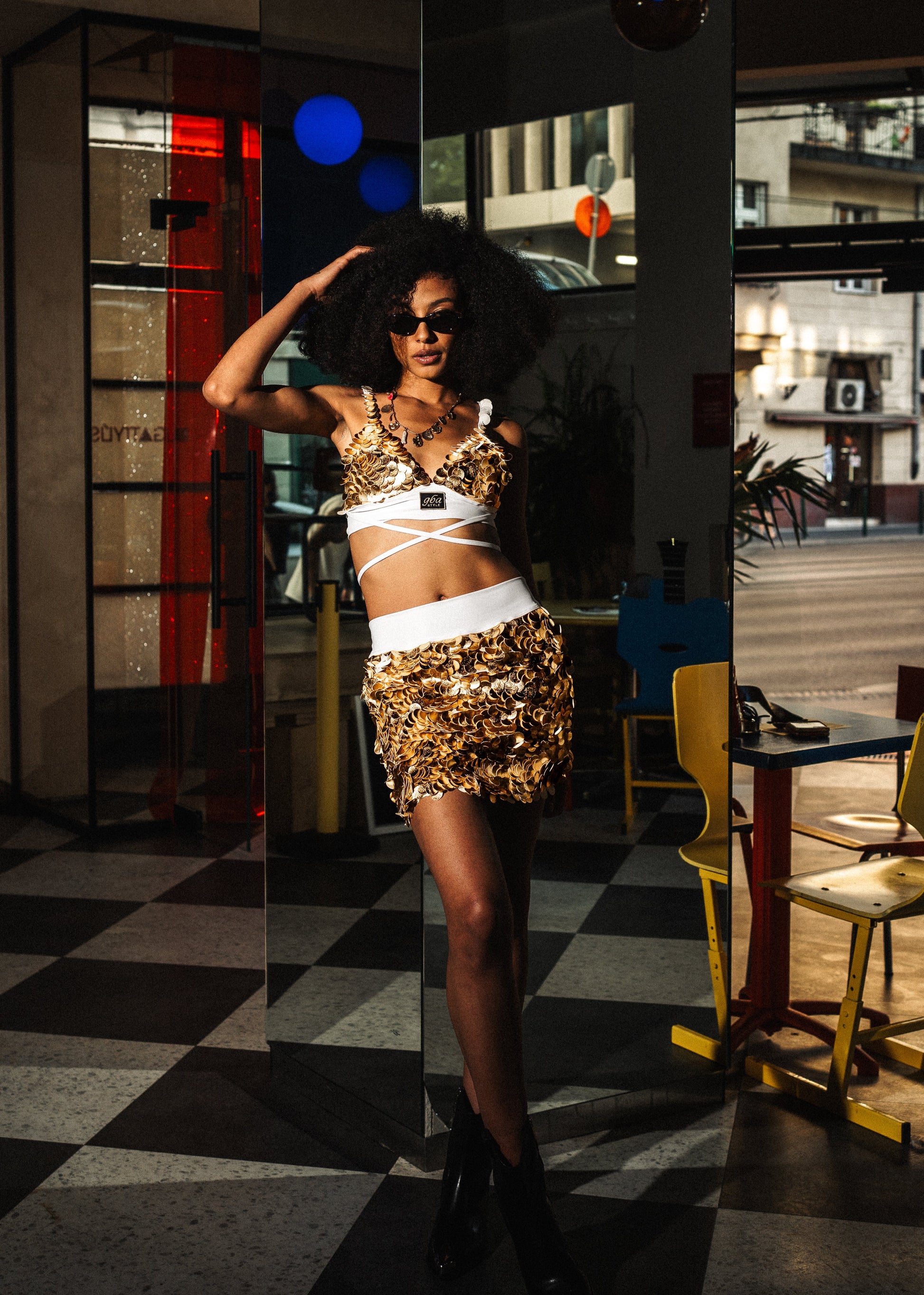 Woman in a gold sequin outfit posing in a room with checkered floor and colorful lights.