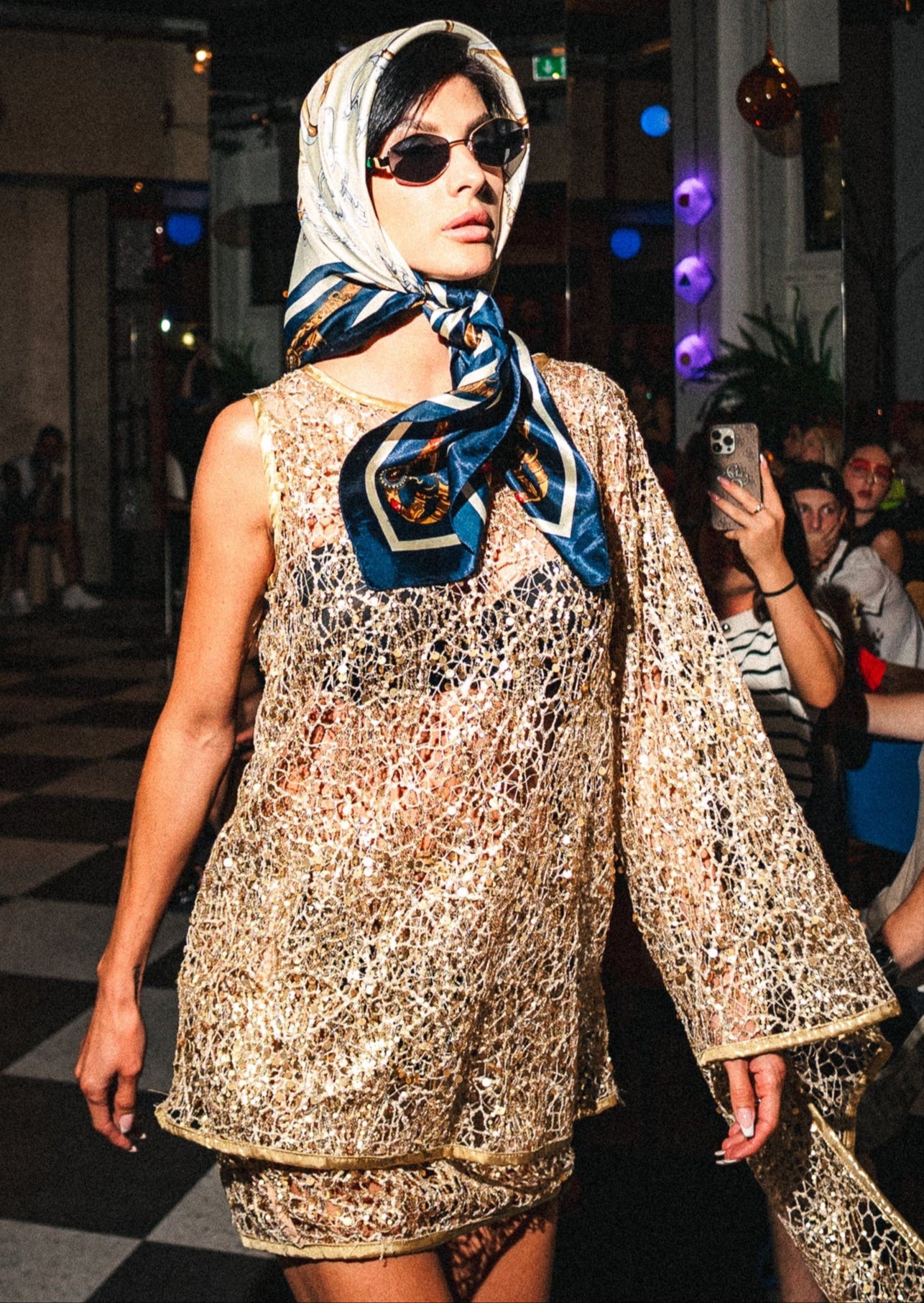 Model walking on a catwalk wearing a patterned dress and headscarf, with an audience in the background.